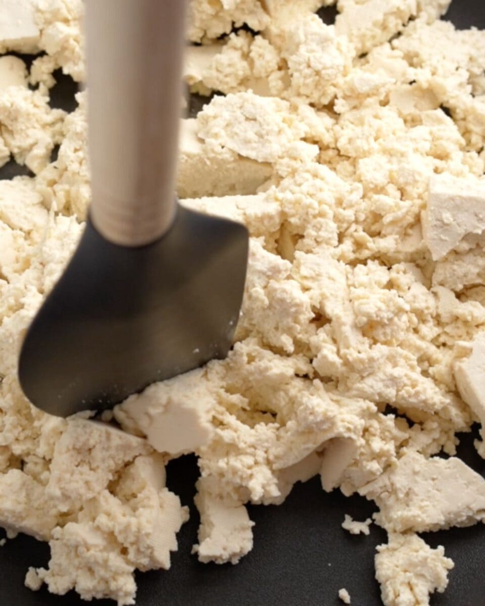 Tofu cooking in a pan with a spatula breaking up big chunks.
