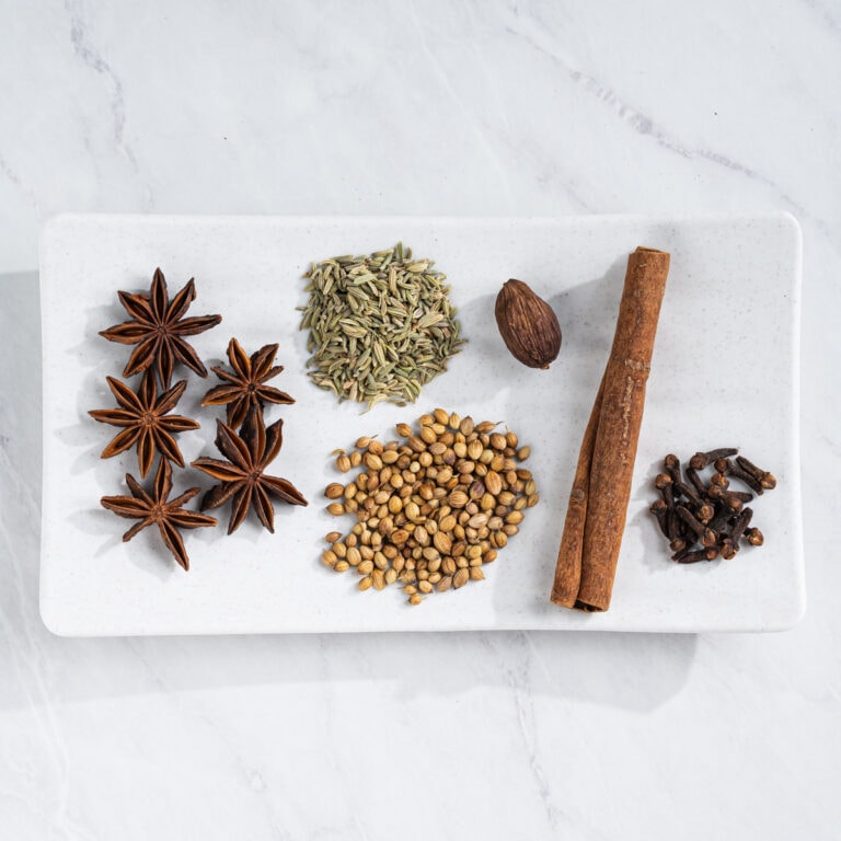 White rectangular plate of pho spices: star anise, fennel seeds, coriander seeds, black cardamom, cinnamon stick, and whole cloves.
