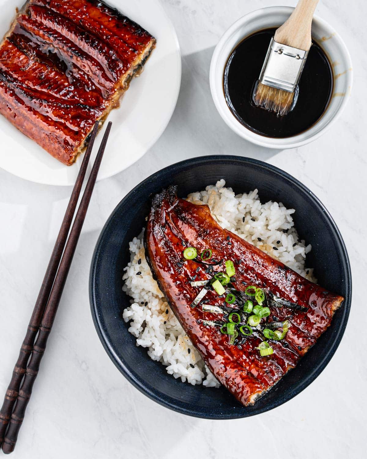 top down of bowl of unagi don with white rice, and grilled eel. Surrounded by plate of grilled eel, bowl eel sauce with sauce brush and a pair of chopsticks.