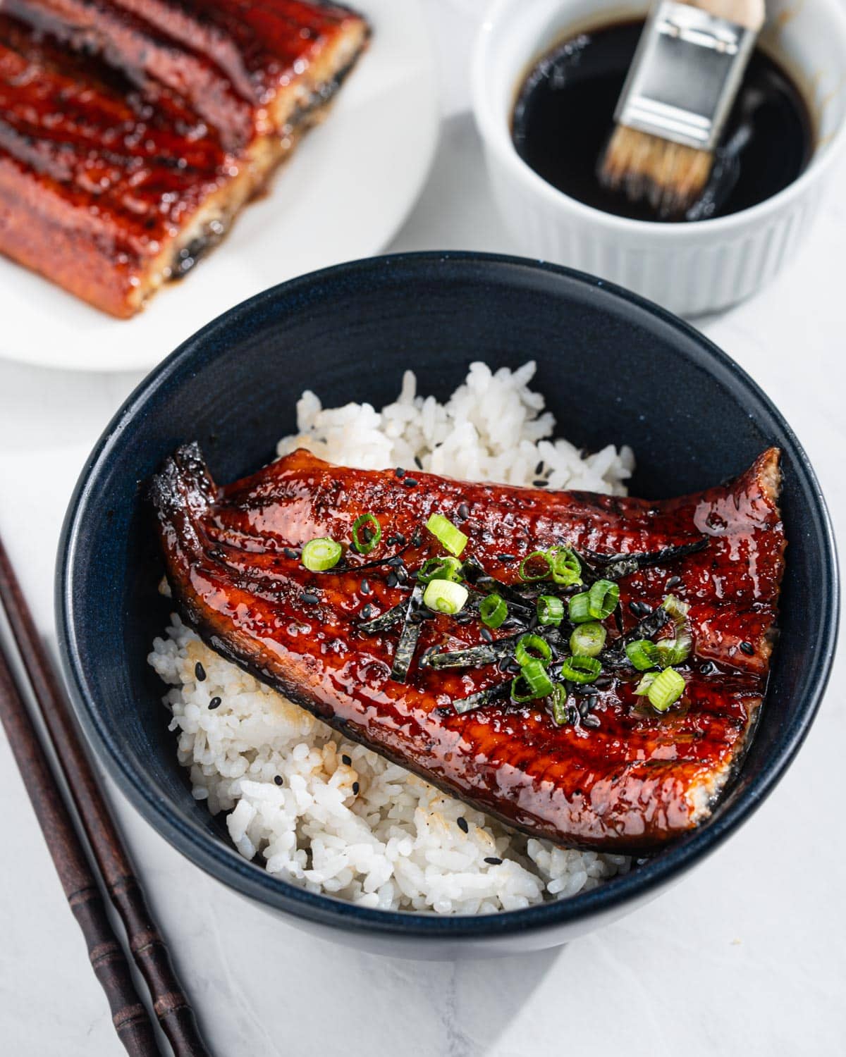 Bowl of unagi don with white rice, and grilled eel. Plate of grilled eel and bowl eel sauce on the side.