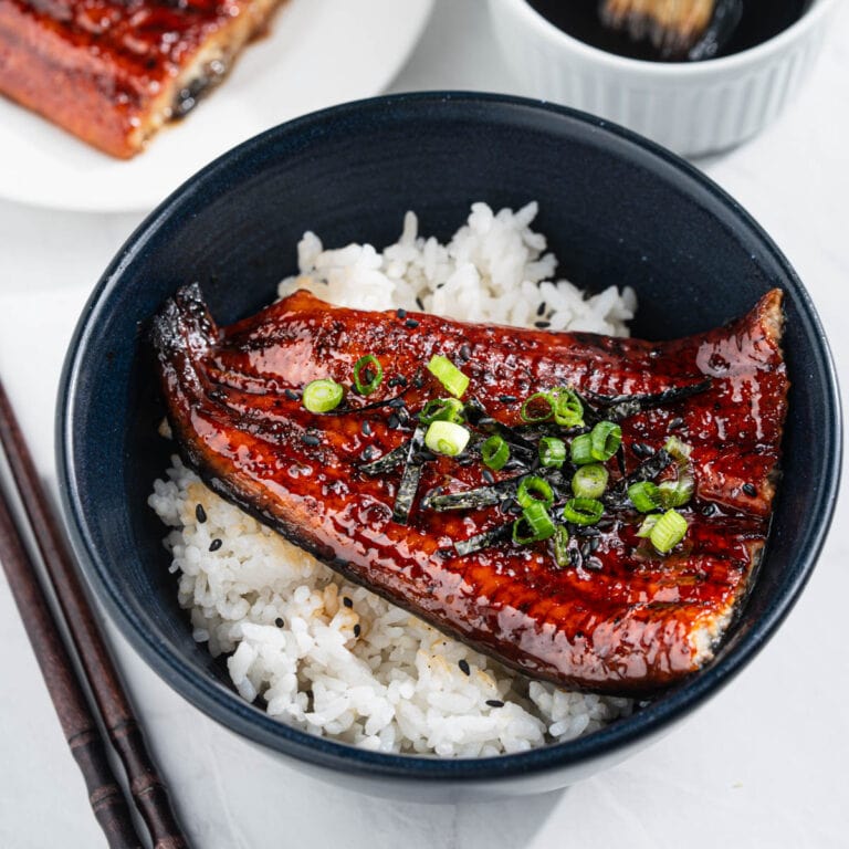bowl of unagi don with white rice, and grilled eel topped with chopped scallions, nori strips, and sesame seeds. Surrounded by plate of grilled eel, bowl eel sauce with sauce brush and a pair of chopsticks.