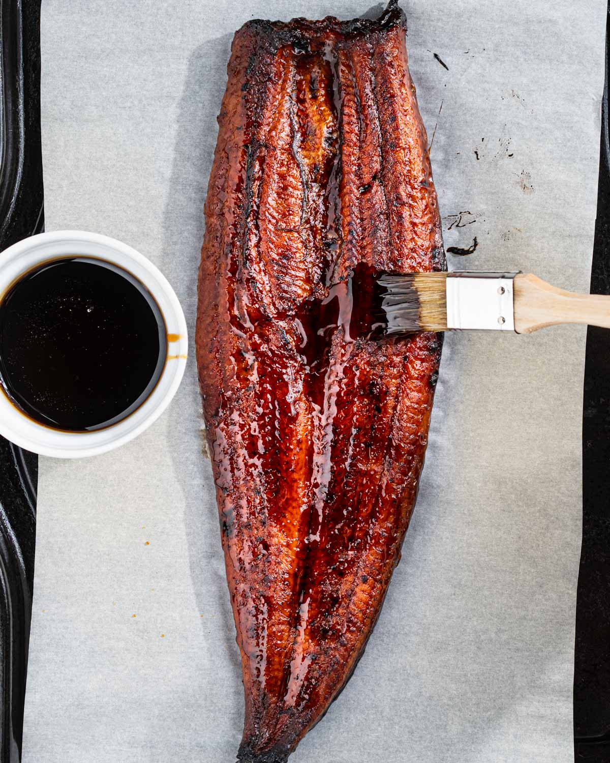 Whole piece of broiled eel on a baking sheet with eel sauce on the side and brush brushing on the sauce.