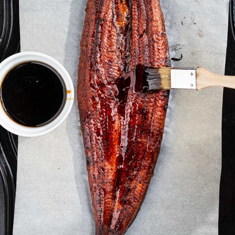 Whole piece of eel on a baking sheet with eel sauce on the side and brush brushing on the sauce.