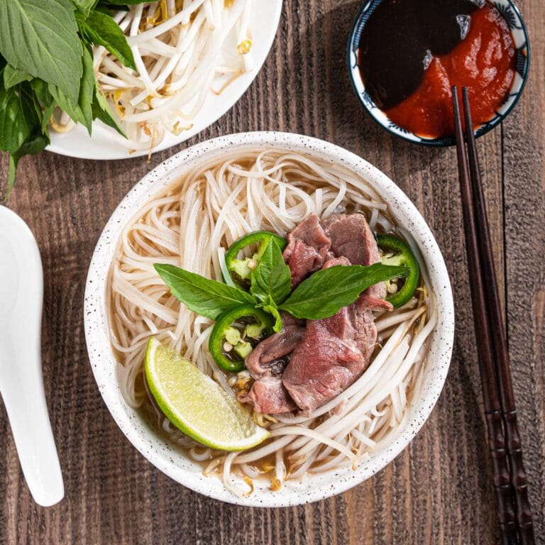 Bowl of pho tai with lime wedges, herbs, hoisin sauce, and sriracha.
