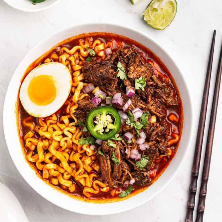 Bowl of beef birria ramen with soft boiled eggs, lime wedges, chopped onions and cilantro.