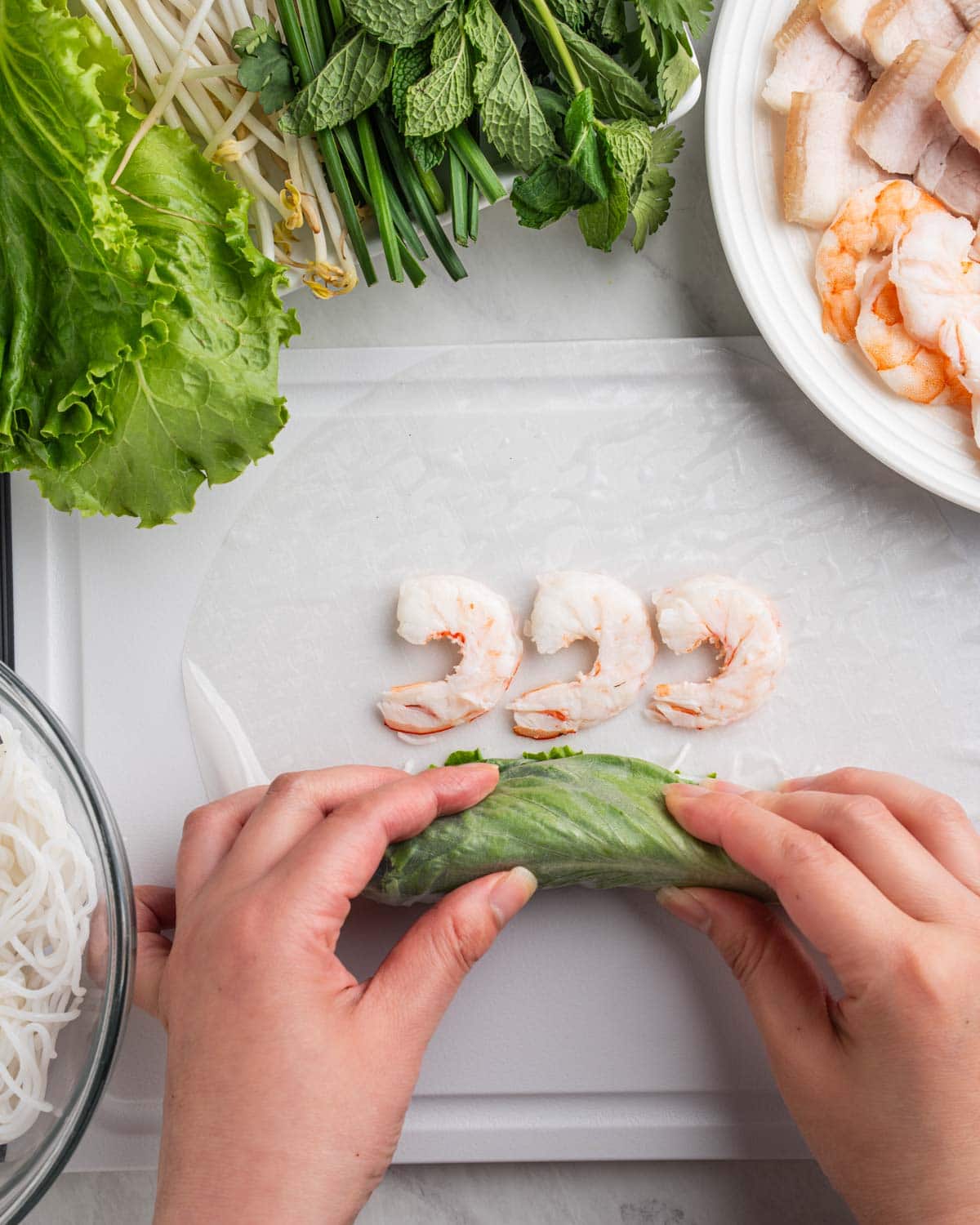 hands tucking in lettuce, herbs and noodles folding towards the shrimp on edge of rice paper.