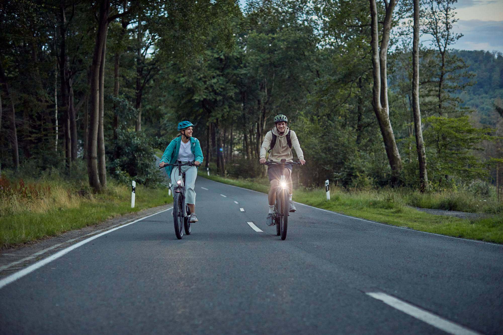 Zwei Radfahrer auf Landstraße in Waldnähe bei Dämmerung.