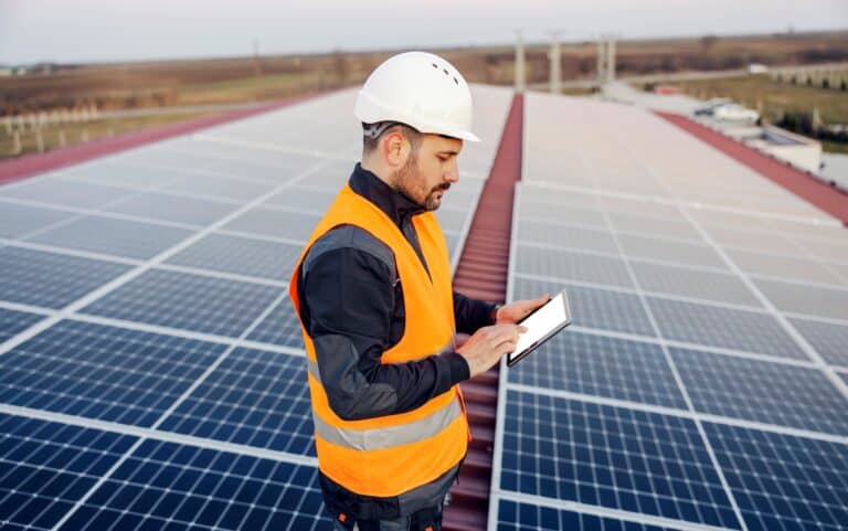 A solar panel technician scrolling on tablet and checking on solar panels on roof