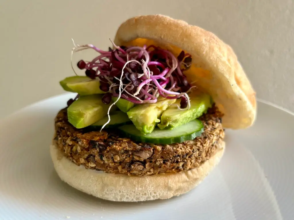A plant-based burger featuring a hearty veggie patty, sliced cucumber, avocado, and vibrant purple sprouts, served in a soft bun on a white plate.