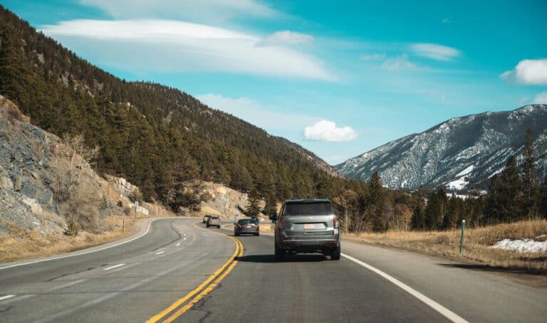 Cars driving on a scenic mountain road during a road trip, with lush green trees and snow-capped mountains in the background.