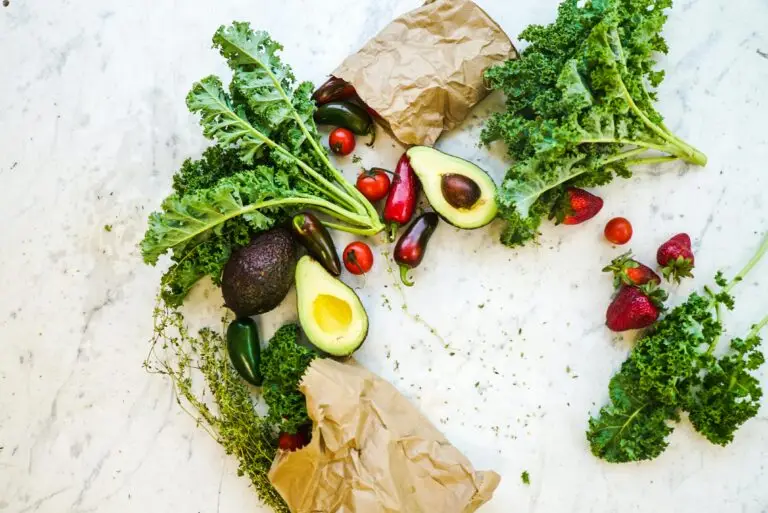 An assortment of fresh vegetables and fruits arranged on a marble surface. The selection includes curly kale, avocados, strawberries, cherry tomatoes, jalapeño peppers, and some sprigs of thyme. A brown paper bag is also visible, partially open and spilling some of the vegetables.