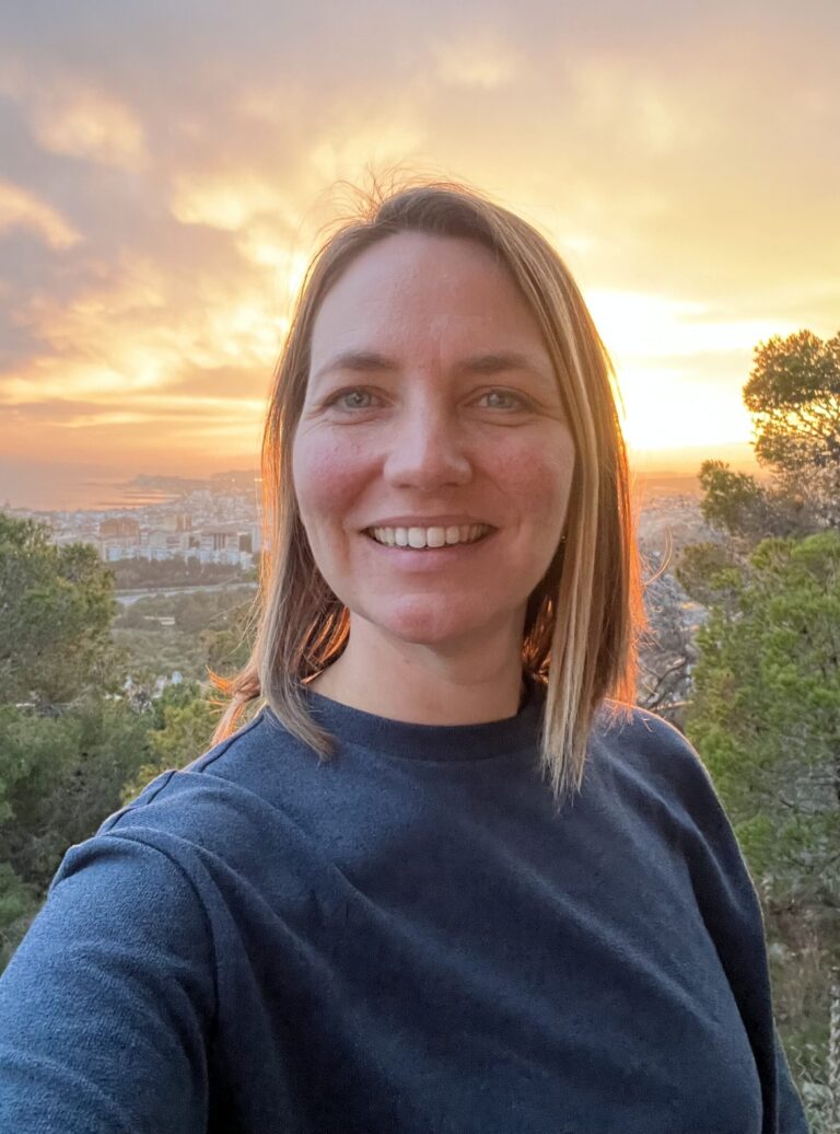 Dr. Sharon Facey with shoulder-length blonde hair, smiling in front of a scenic sunset view, with trees and a city visible in the background.
