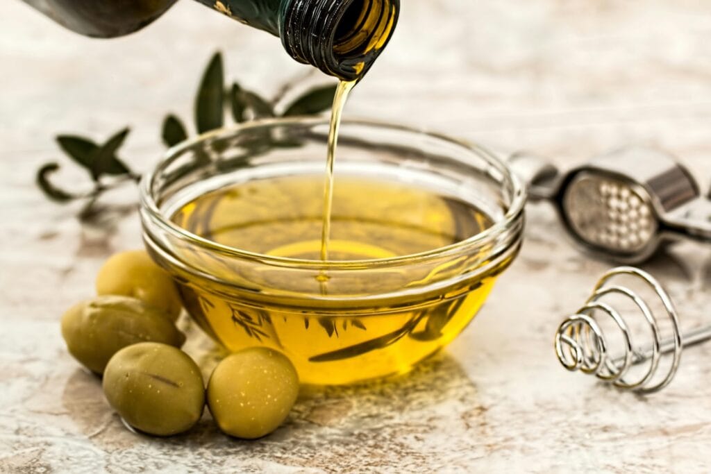 Olive oil being poured into a glass bowl next to green olives and kitchen utensils, illustrating healthy cooking choices