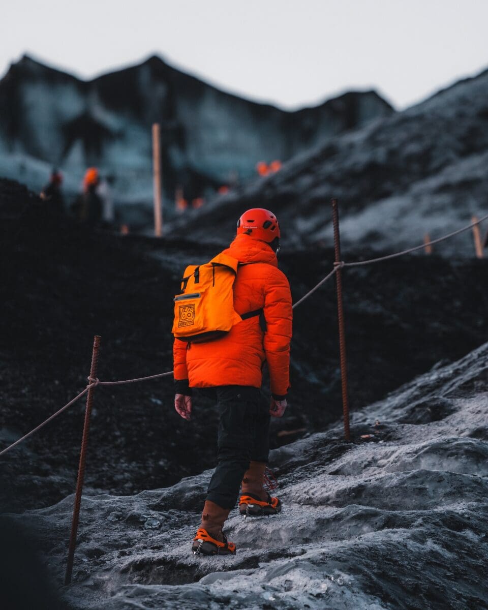 A man wearing a red jacket and an orange backpack turning our back to us walking on a Katla Ice Cave tour. Icy mountains in the background that are black.