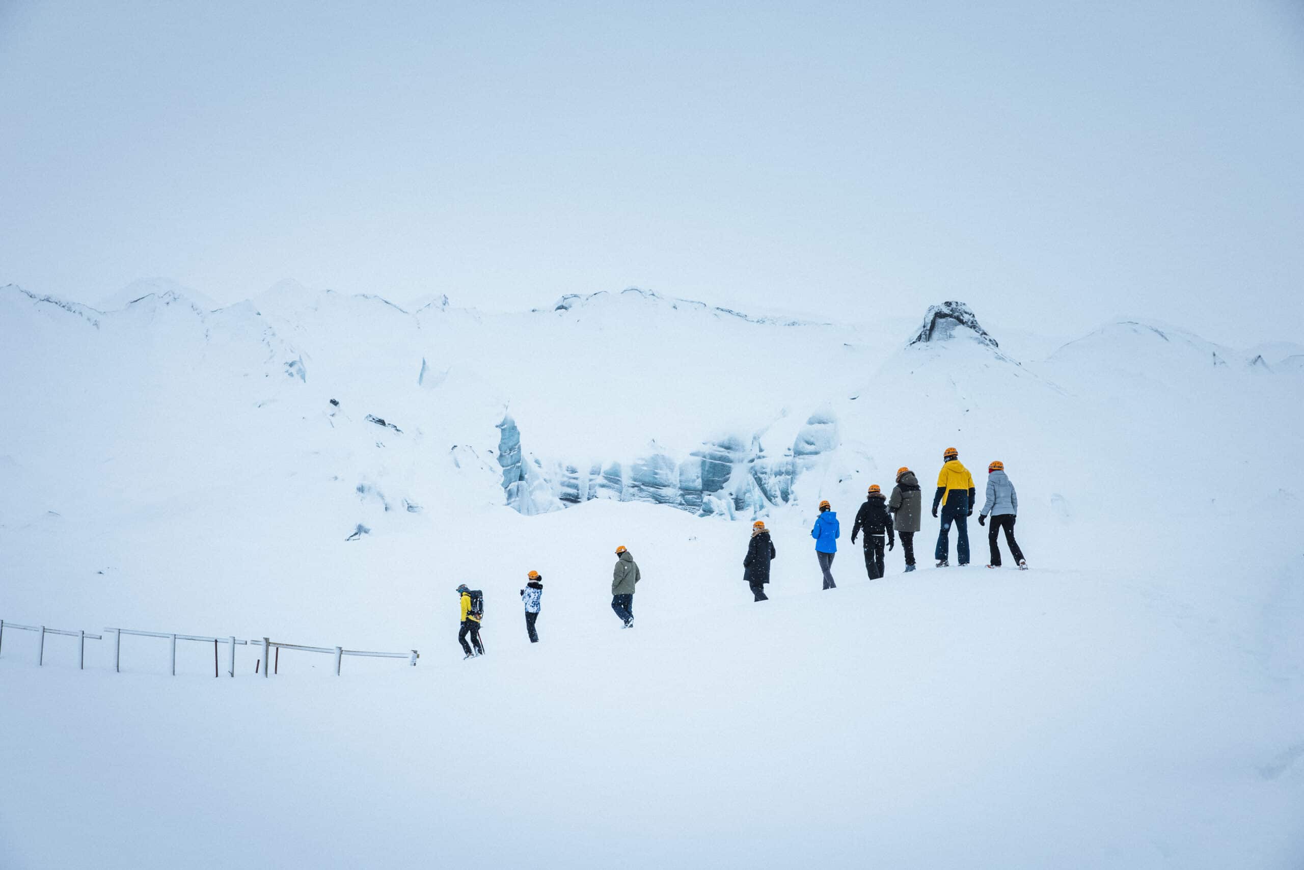 People with orange helmets walking down a hill that is covered in snow and ice. A mountain behind them with white show from Katla Ice Cave.