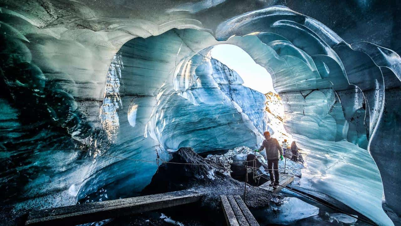 An Ice Cave with icy walls with a circular opening and a man standing on a small bridge walking inside the cave.