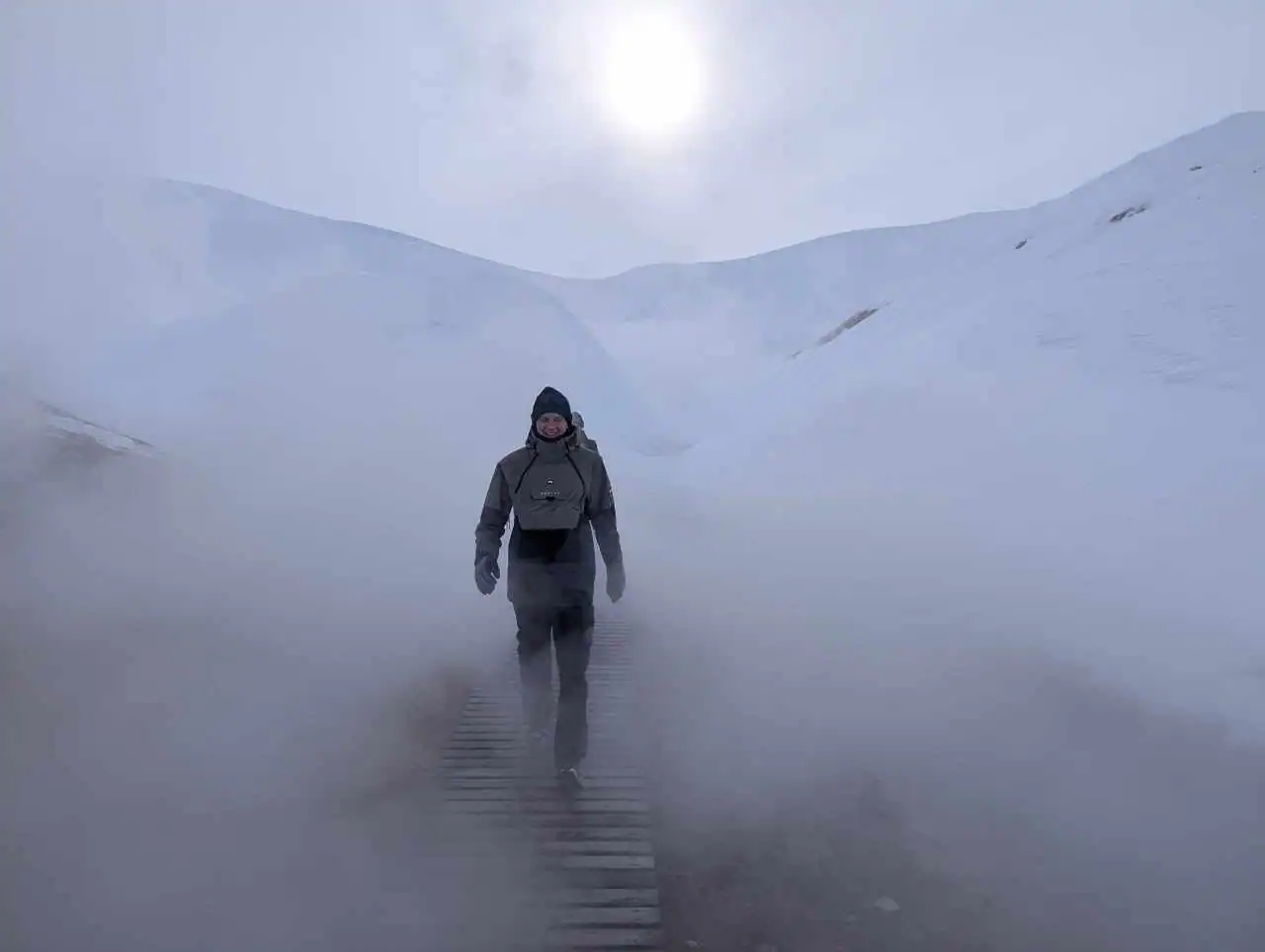A man walking on a wooden trail surrounded by snowy mountains and a mist in the icelandic nature