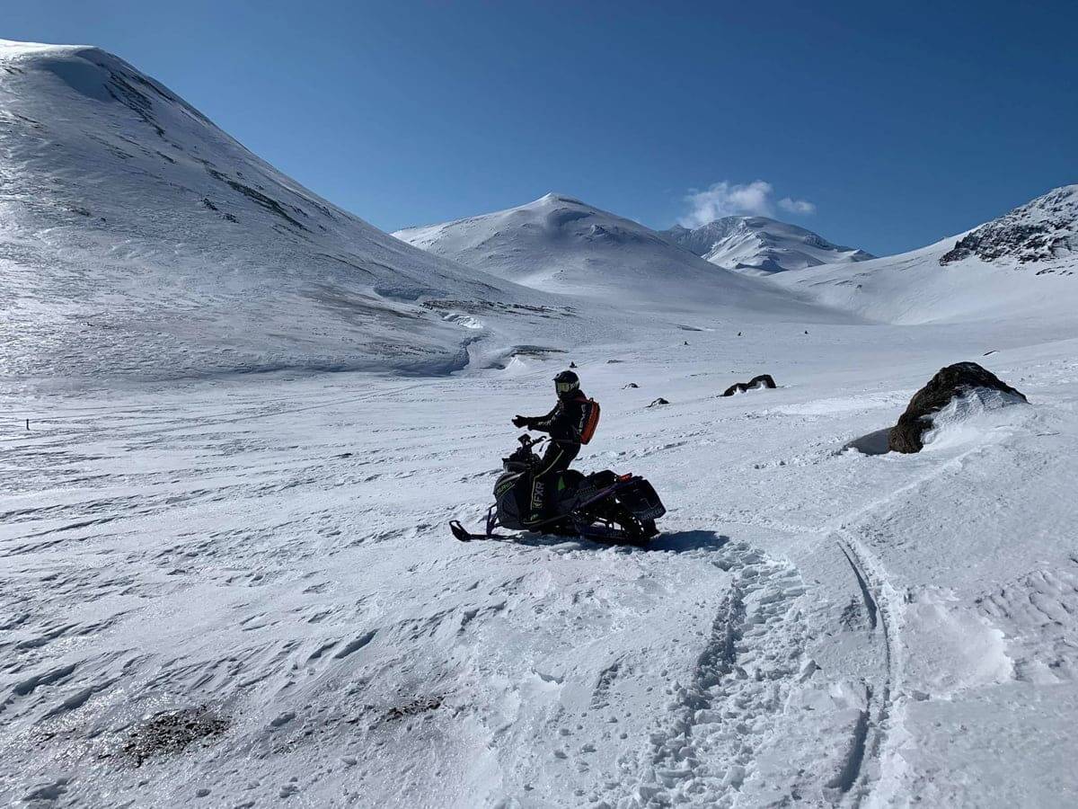 A person on a snowmobile on snowy ground with snowy mountains in the back with clear blue sky on an adventure tour with Southcoast Adventure in Iceland.
