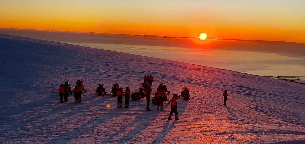 Multiple people on a snowmobile on Eyjafjallajokull highland watching a sunset where the sky is orange and the orange sun shines on icy snow.