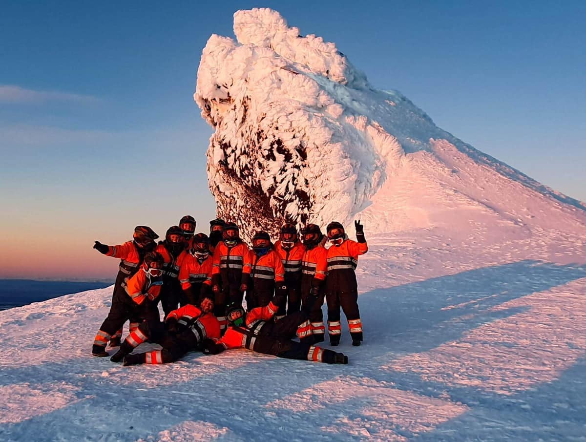 Multiple people standing with a large ice behind them wearing orange and black snowsuites and helmets from a snowmobiling tour with Southcoast Adventure on Eyjafjallajokull