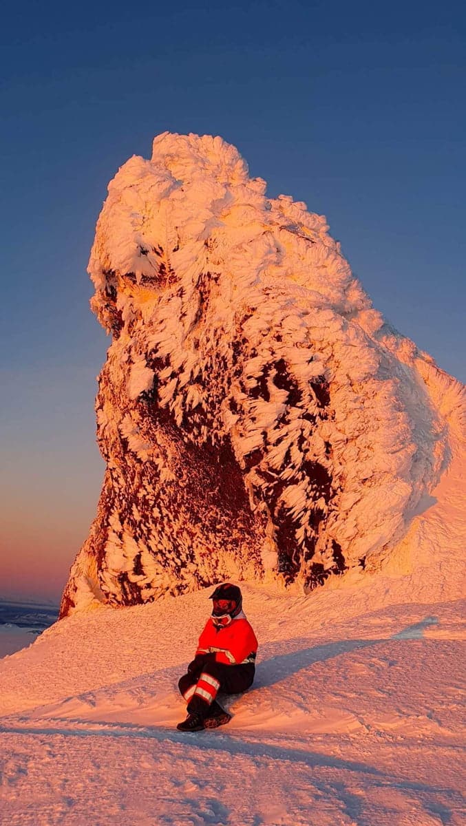 A person wearing an orange and black snowsuit and a helmet sitting on snowy ice with a large snowy ice behind her with orange light from a sunset on Eyjafjallajokull tour with Southcoast Adventure.