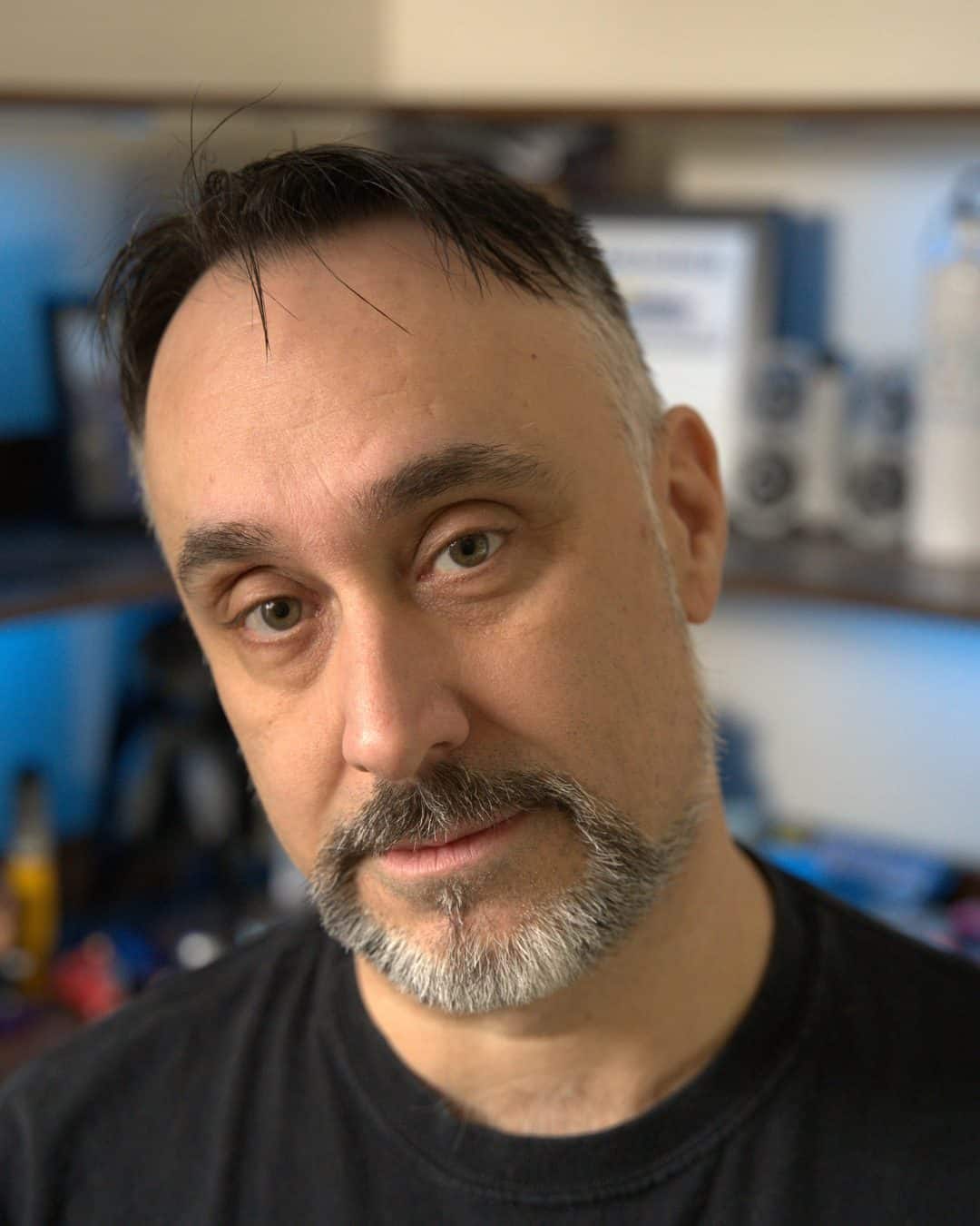 Casual portrait of Glenn Murano, founder of Southington Digital Solutions, in a black t-shirt with a neutral expression, photographed in a workspace with blurred shelves and tech equipment in the background.
