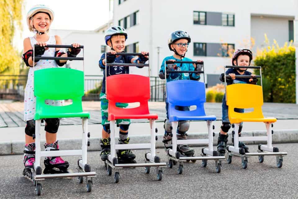 Kinder auf Rollstühlen mit bunten Sitzflächen vor moderner Wohnanlage.