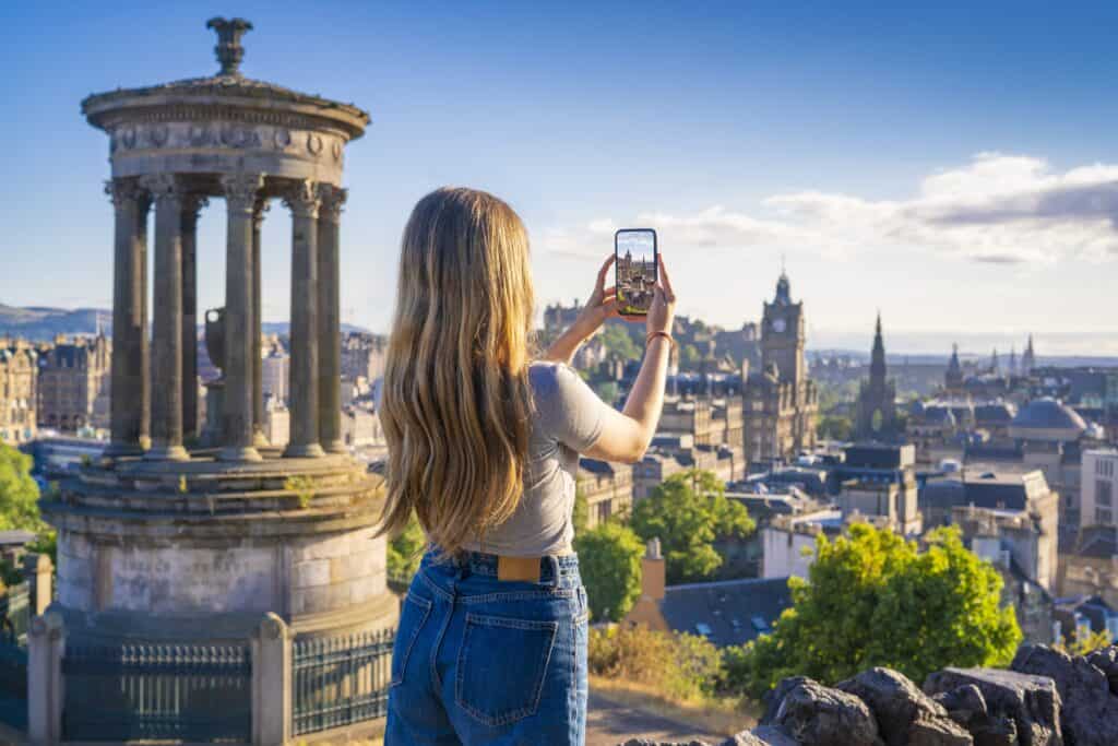 City view from Edinburgh rooftop with historic landmarks and a woman taking a photo.
