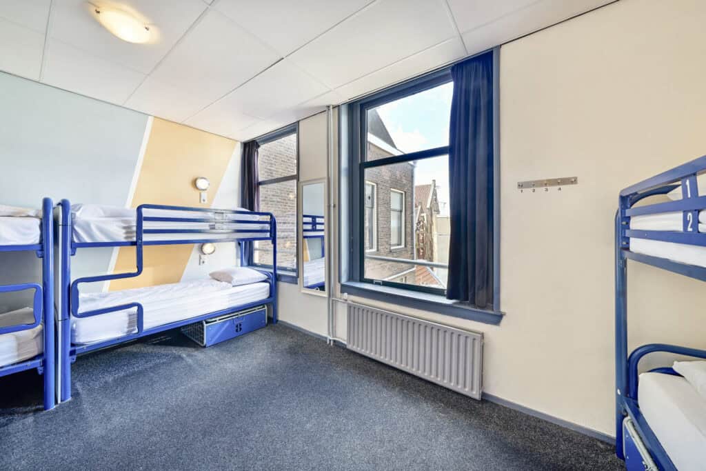 Hostel dorm room featuring blue bunk beds, large windows with city views, and a cosy, clean atmosphe.