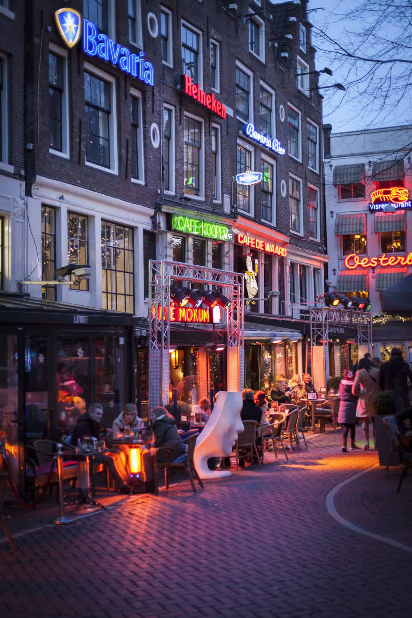 Vibrant street scene outside The Flying Pig Downtown hostel with cafes and bars in Amsterdam at dusk.