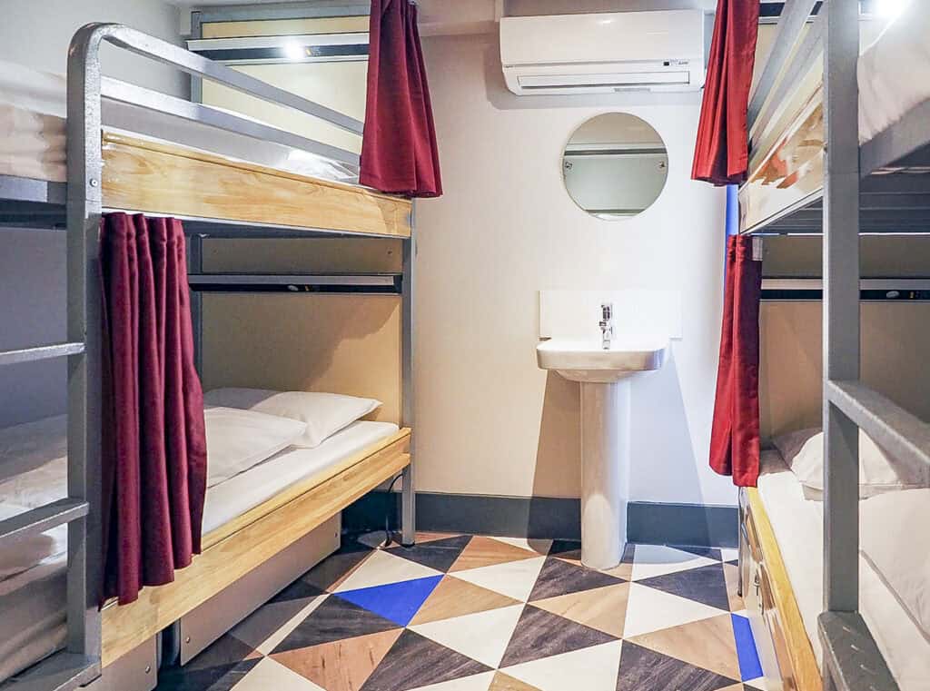 Hostel dorm room with bunk beds, privacy curtains, and a small sink.
