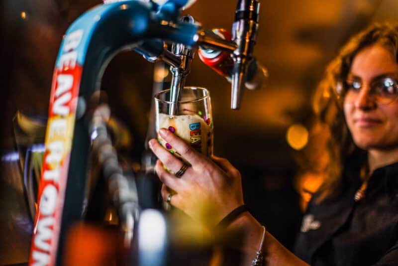 Woman pouring beer from tap at Liverpool Street Hostel bar, lively atmosphere.