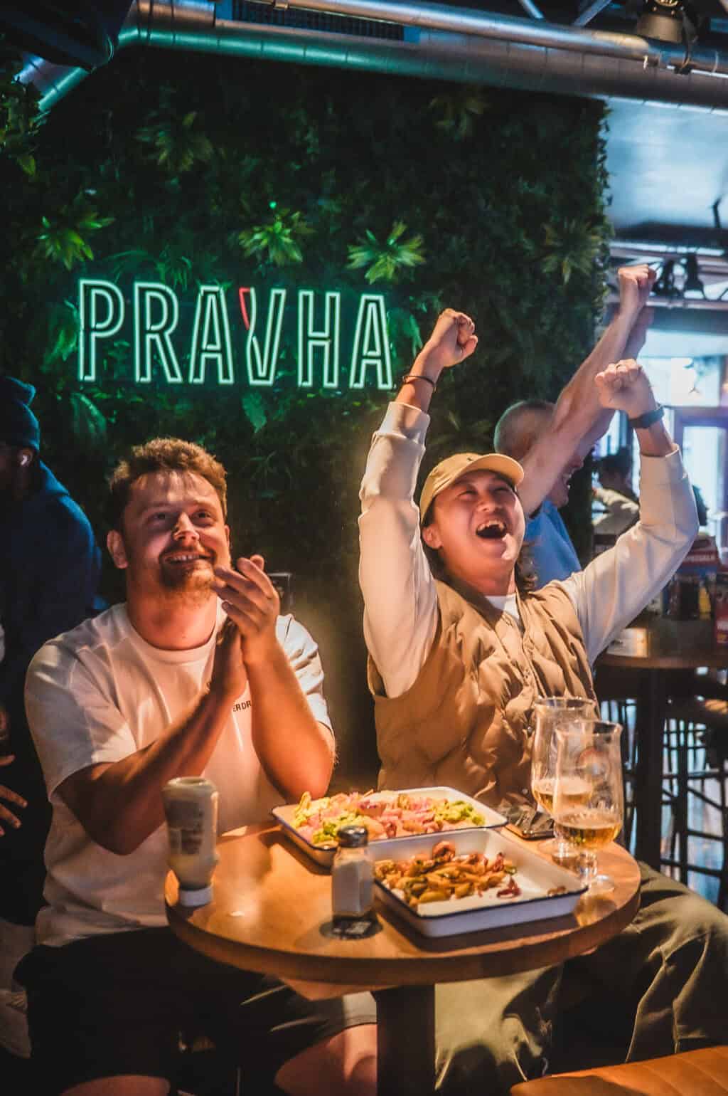 Two friends enjoying drinks and food at a Prague-themed bar with a neon 