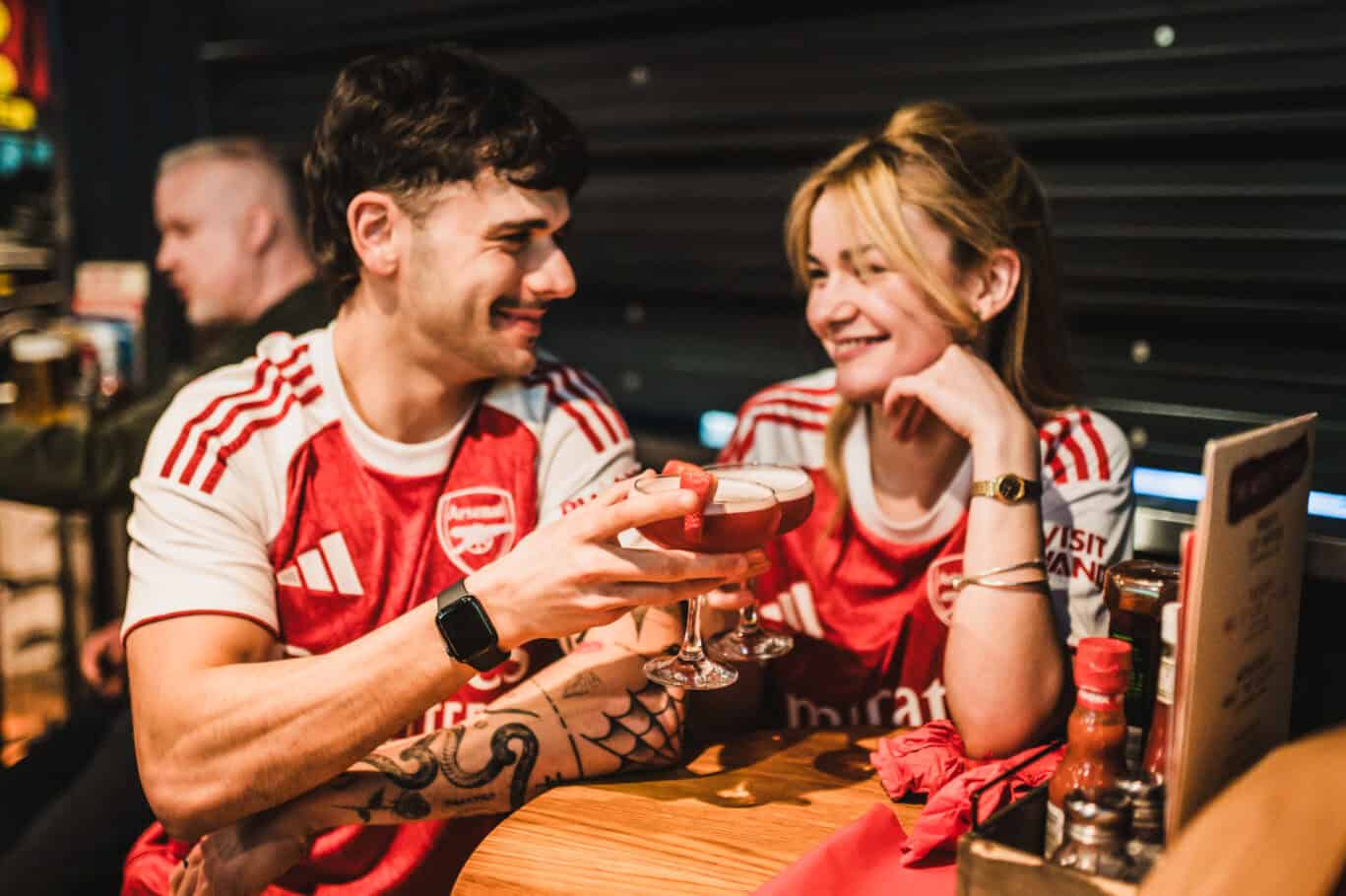 Two friends in Arsenal football shirts enjoying drinks at London Bridge Hostel bar.