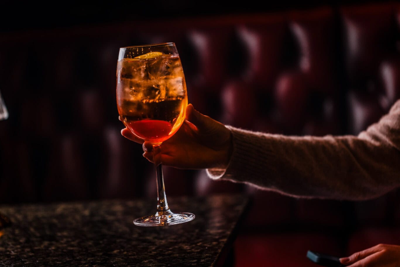 A person holding a glass of orange cocktail in a dimly lit bar setting.