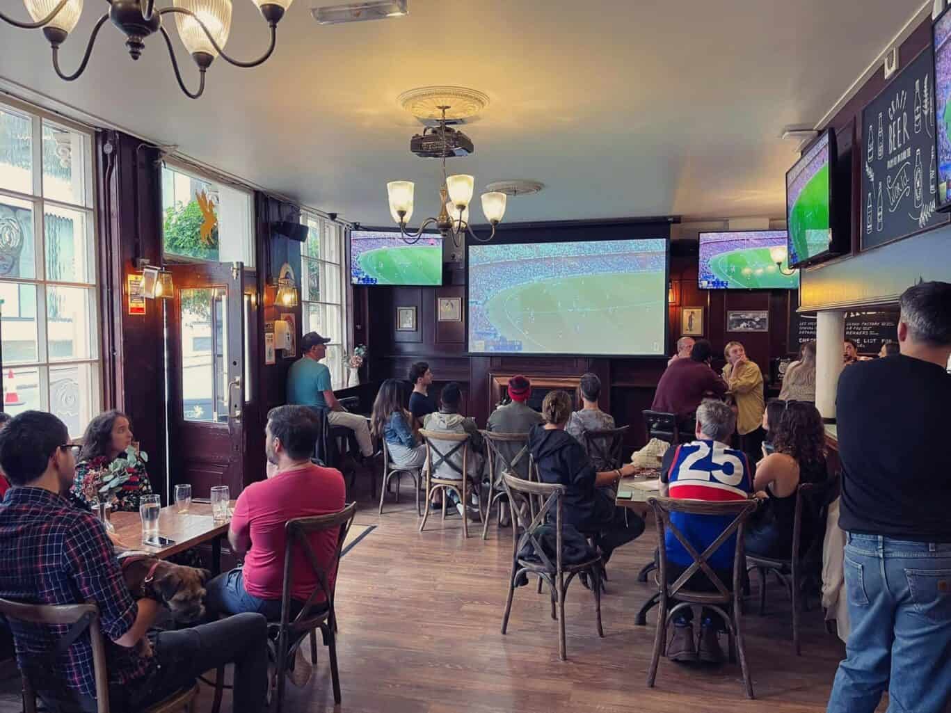 Guests watching a football match in a lively hostel lounge with multiple screens.