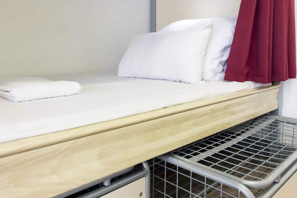 Single bed in hostel room with white bedding and red curtain.