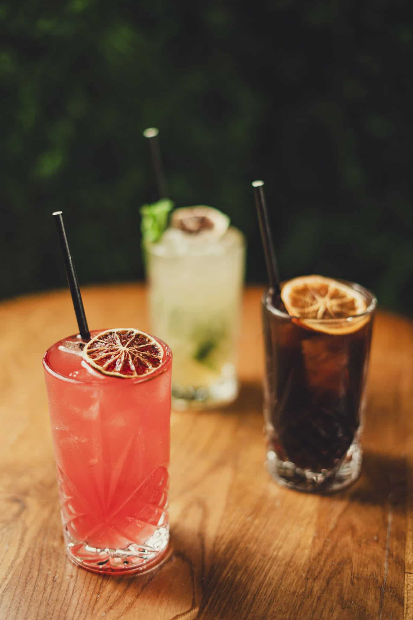 Three colourful cocktails with citrus slices on a wooden table at Shepherd's Bush Hostel.