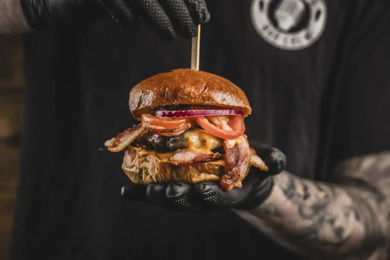 Close-up of a person holding a gourmet burger with bacon, cheese, and vegetables.