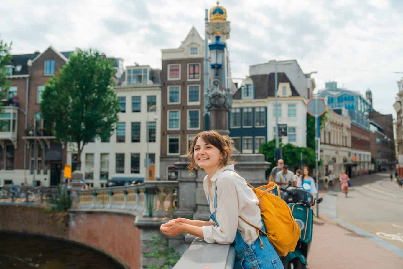Young woman with backpack enjoying Amsterdam canal view, vibrant cityscape background.