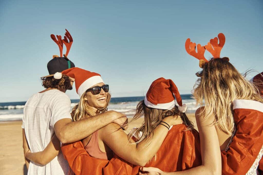 Group of friends celebrating Christmas on the beach, wearing Santa hats and reindeer antlers, enjoyi.