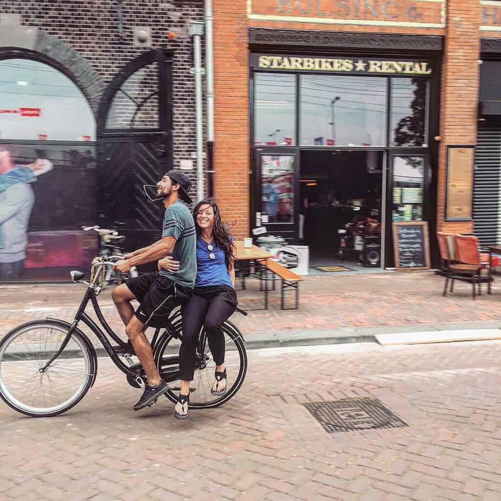 Young couple riding a bicycle in front of Mitte Hostel Berlin, vibrant city scene.