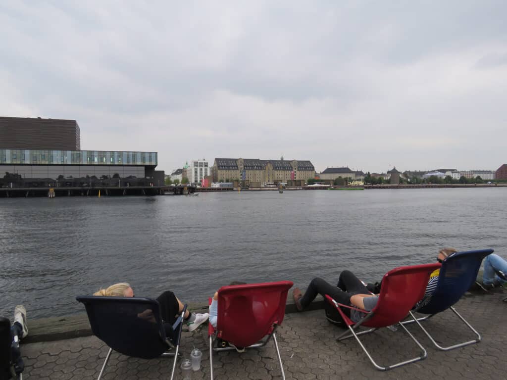 Riverside view of Mitte Hostel Berlin with people relaxing by the Spree River. Modern cityscape and.