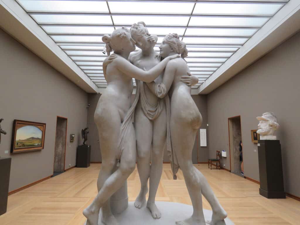 Ancient Greek sculpture of three women embracing, displayed in a modern art gallery.
