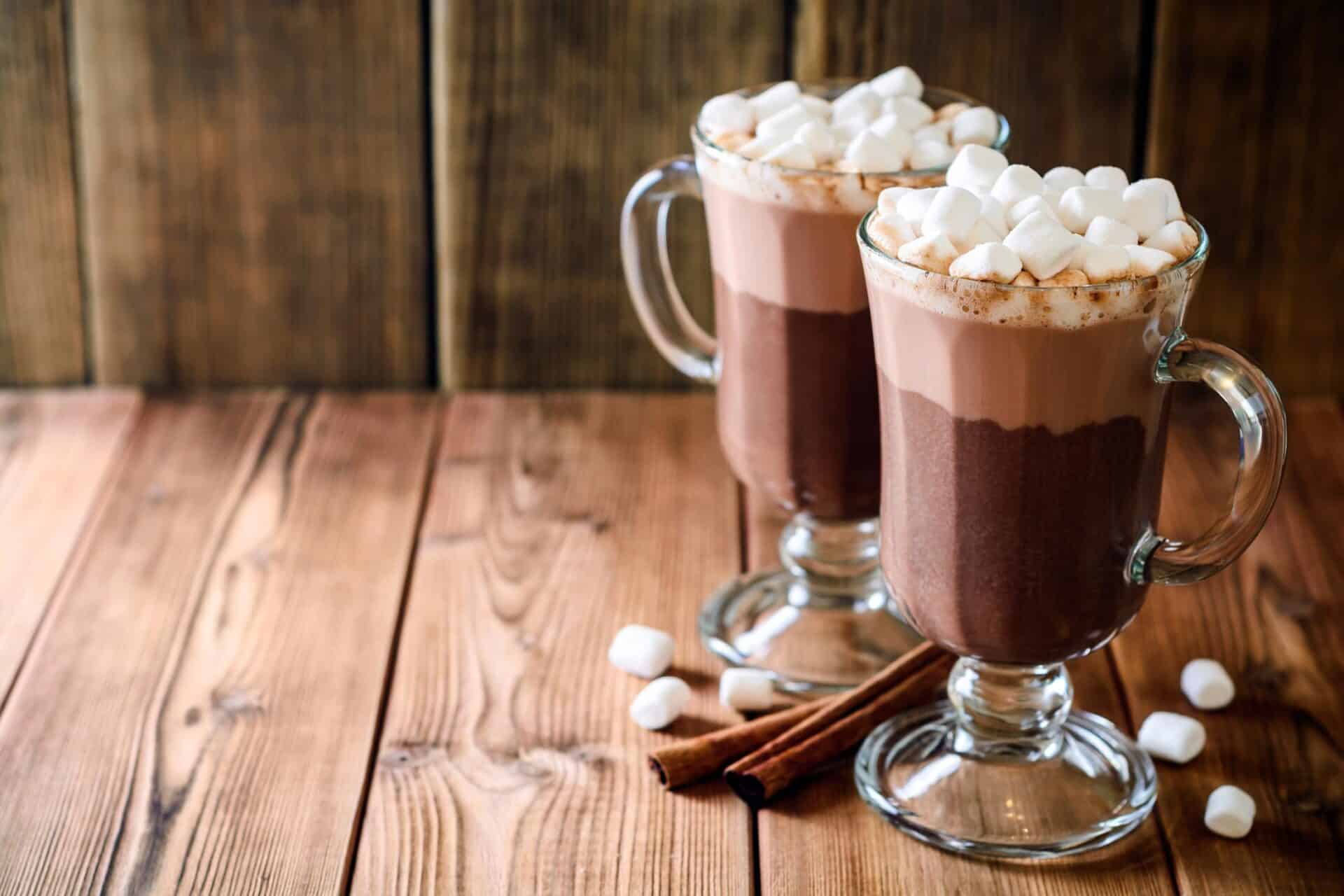 Chocolate hot chocolate with marshmallows in glass mugs, cinnamon sticks on wooden table.