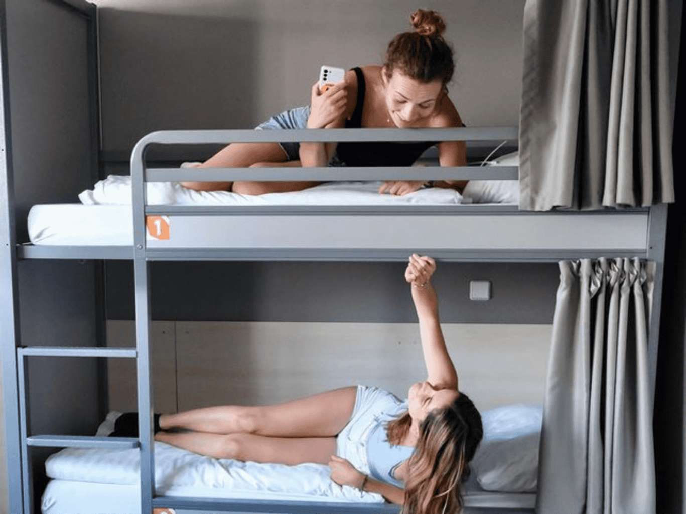 Young woman on top bunk talking on phone, girl on bottom bunk reaching up, hostel room scene.