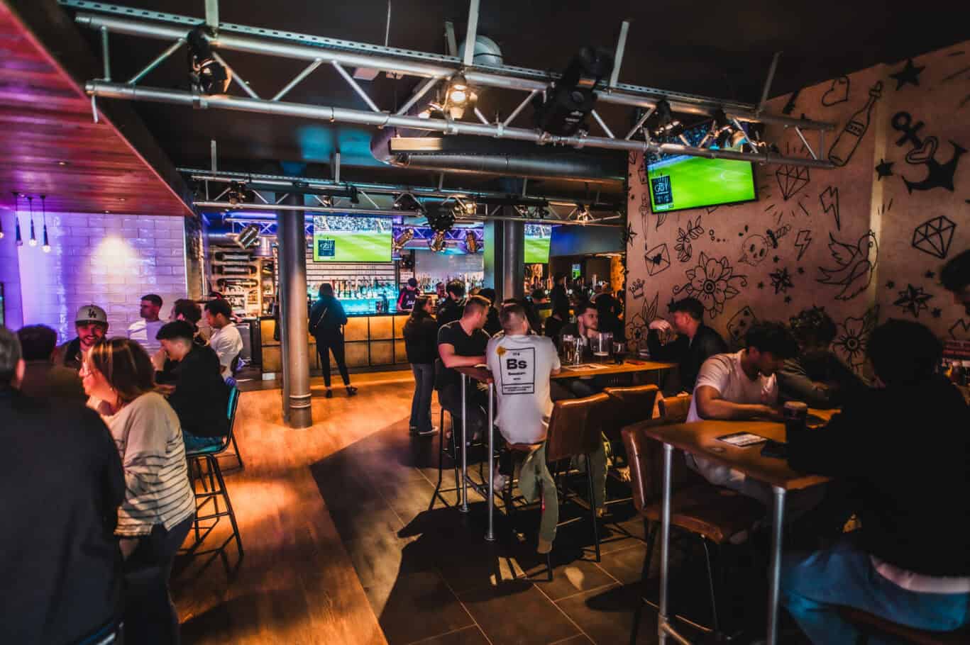 A lively social hostel bar with multiple screens showing sports and groups of travellers enjoying a.