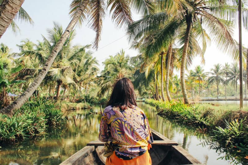 A woman sitting on a boat in a lush, tropical canal with palm trees, representing a relaxing hostel.