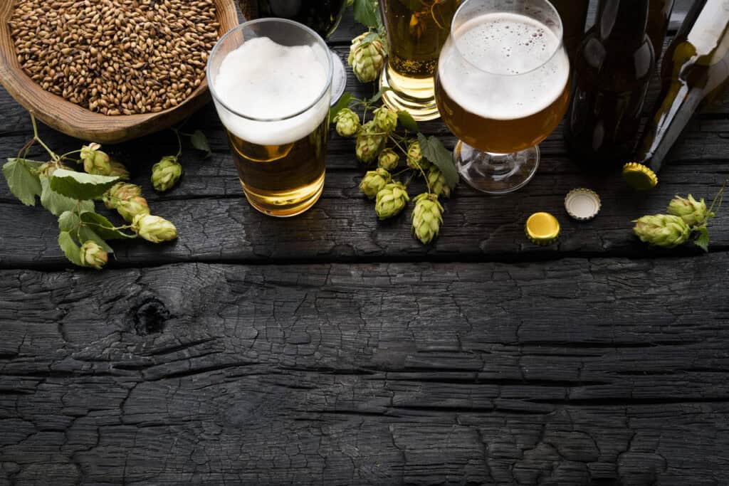 Three glasses of beer with hops and brewing ingredients on a rustic wooden table.