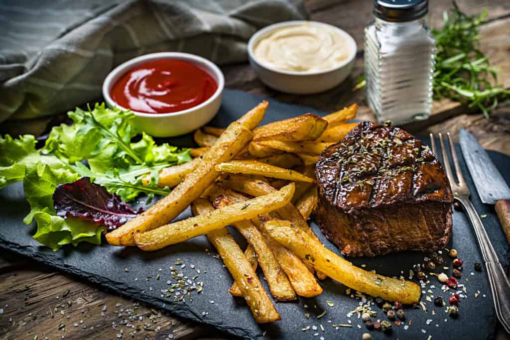Juicy grilled steak with crispy fries, fresh salad, and condiments on a rustic wooden table.